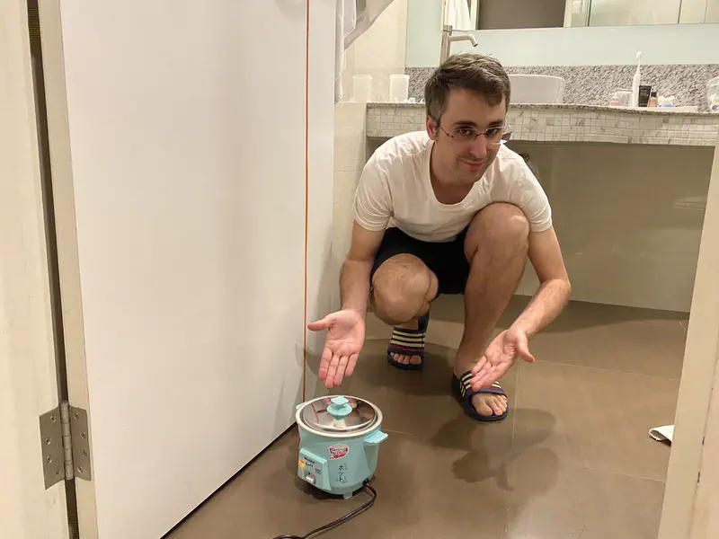 Man crouching on the floor in a bathroom, presenting a small turquoise rice cooker.
