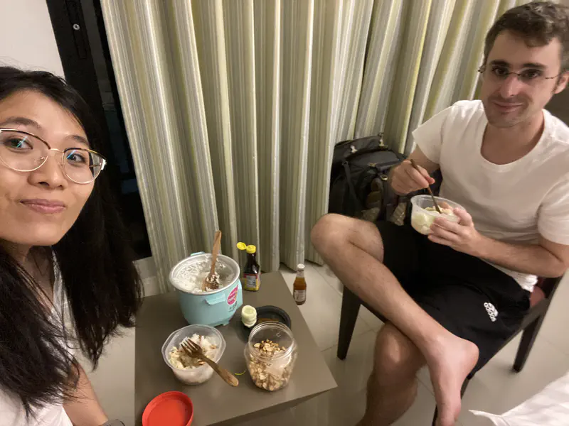 Two people eating rice from bowls inside a room, with a small turquoise rice cooker and condiments on the table.
