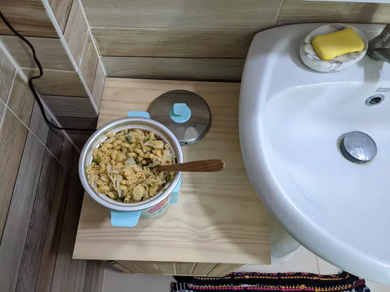 Small turquoise rice cooker on a wooden shelf by a sink, filled with macaroni and vegetables.