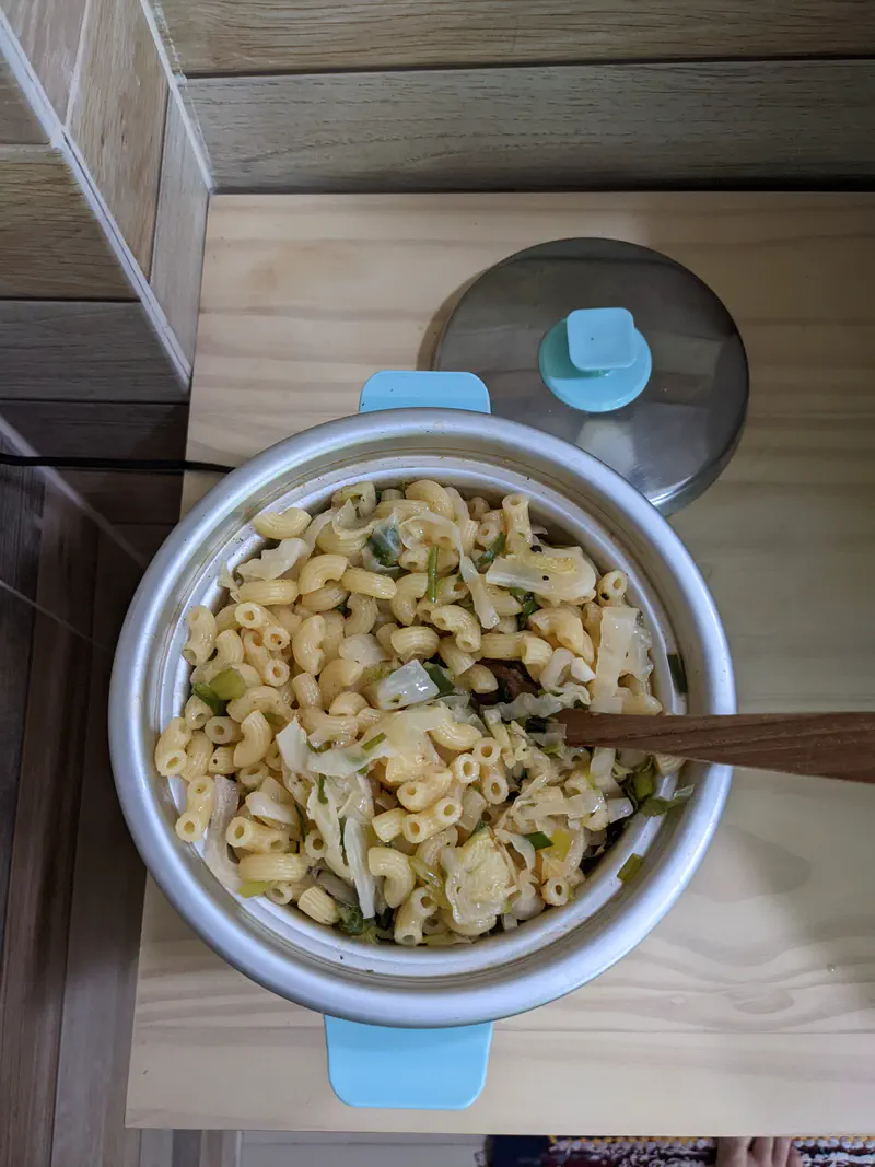 Close-up of macaroni with vegetables in a small turquoise rice cooker with a wooden spoon inside.