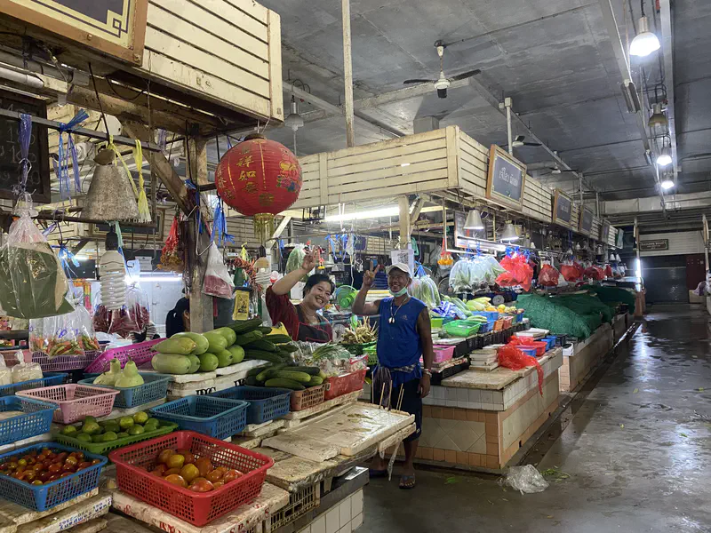 Vendors smiling and waving at a fresh market stall filled with vegetables and fruits displayed in baskets.