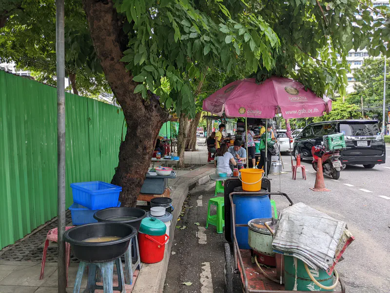 Street food stall under a pink umbrella with people eating at small tables by the roadside.