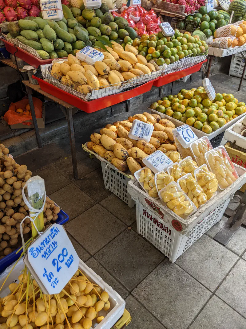 Fruit market stall displaying mangoes, oranges, longans, and jackfruit neatly arranged in baskets and trays with price signs.