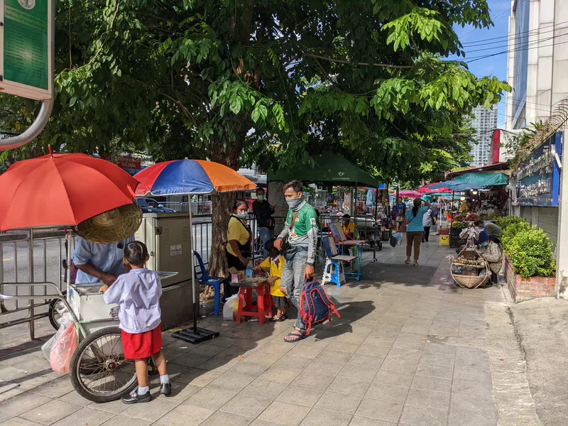 Sidewalk with street vendors under colorful umbrellas selling food and goods, with people walking and shopping.
