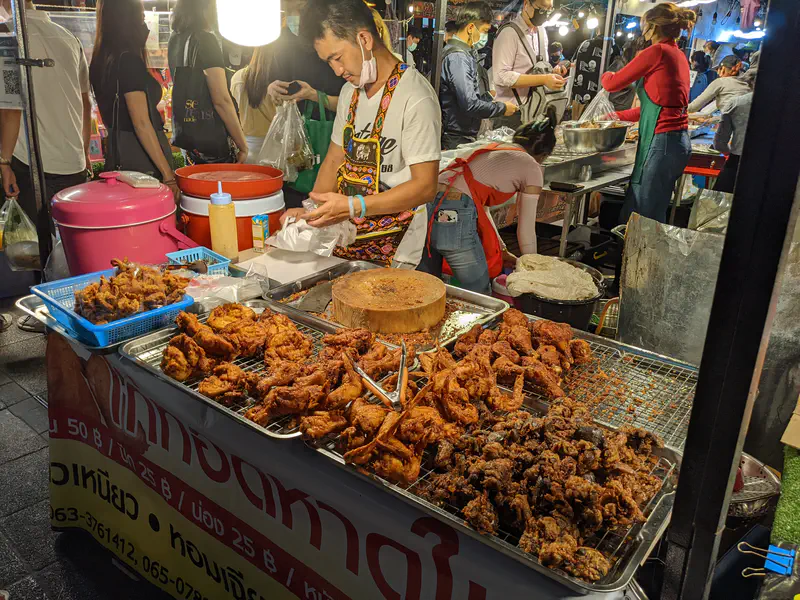 Vendor at a busy night market stall selling trays of fried chicken pieces while preparing food for customers.