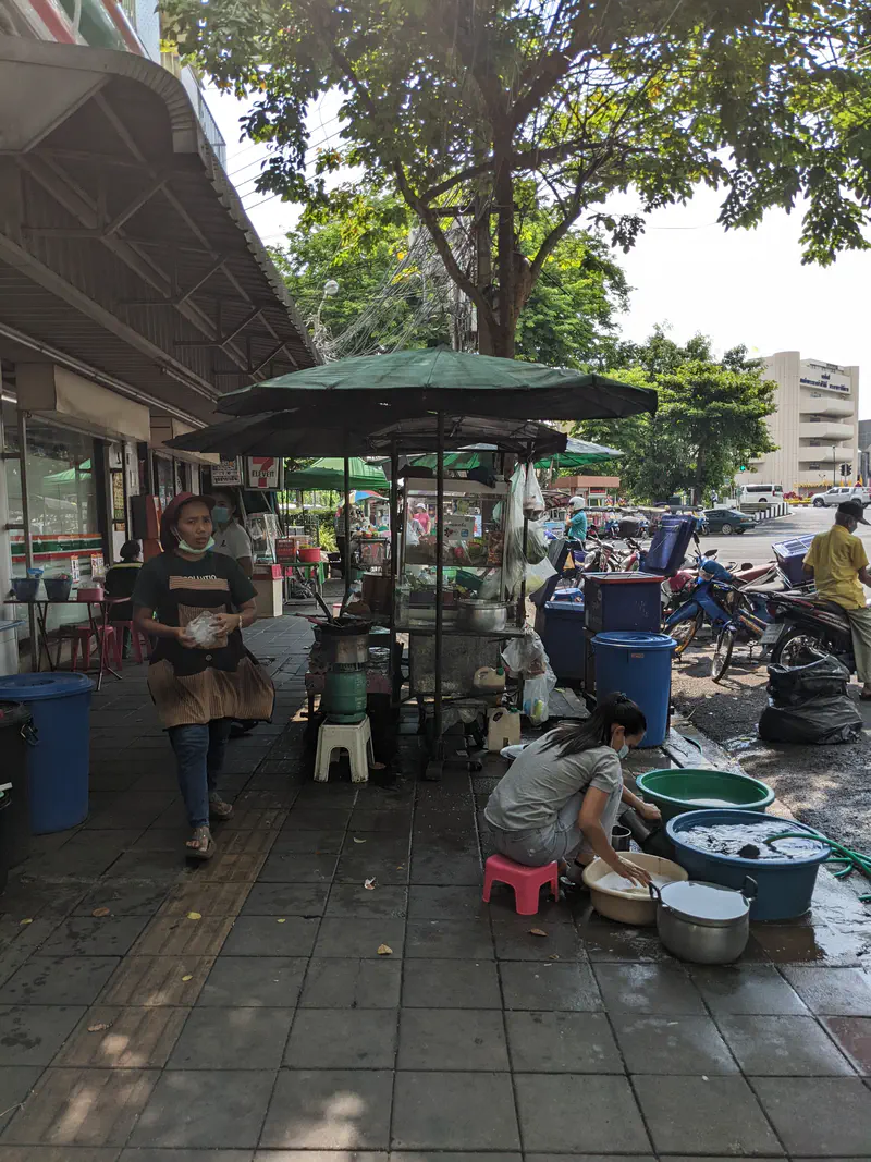 Street vendor washing dishes on the sidewalk next to a food stall under a green umbrella.