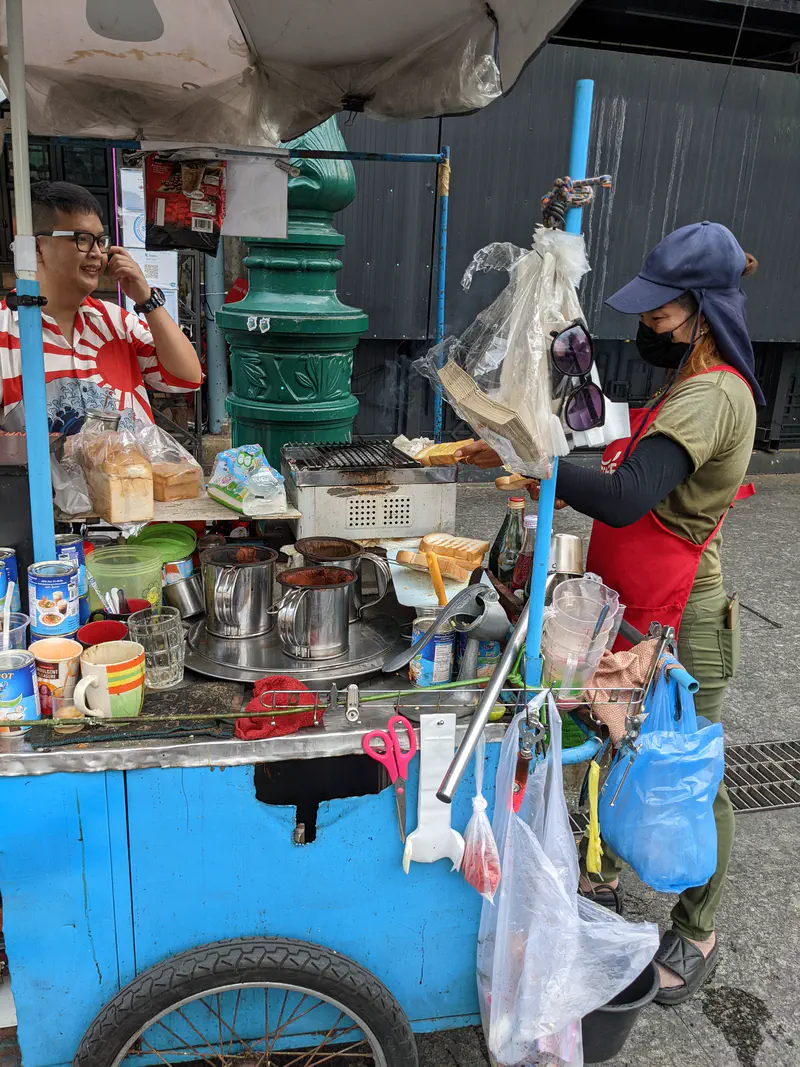 Small street cart with mugs, pots, and bread where a vendor prepares food and drinks while a customer smiles.