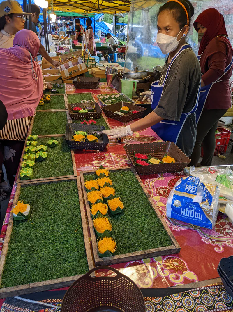 Market vendor arranging small Thai desserts in green banana leaf cups on display trays.