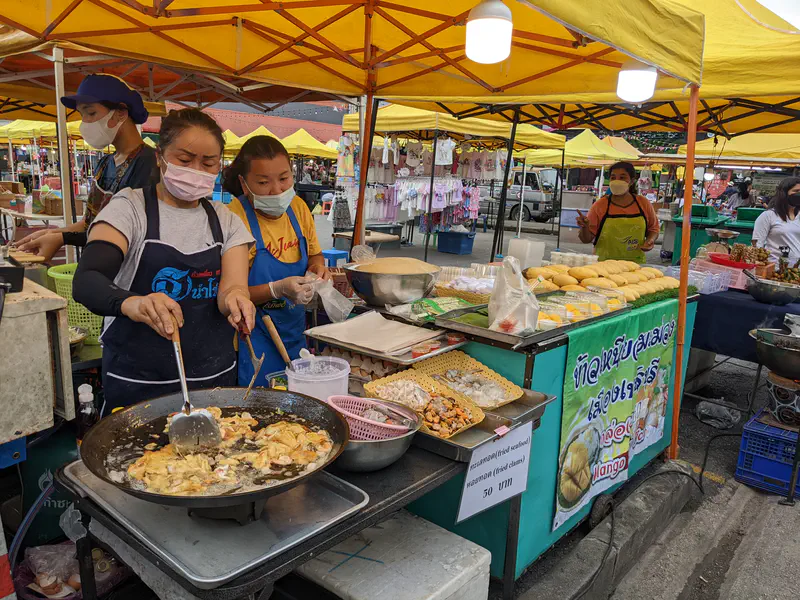 Vendors frying seafood in a large wok at a busy market stall with trays of fresh mangoes in the background.