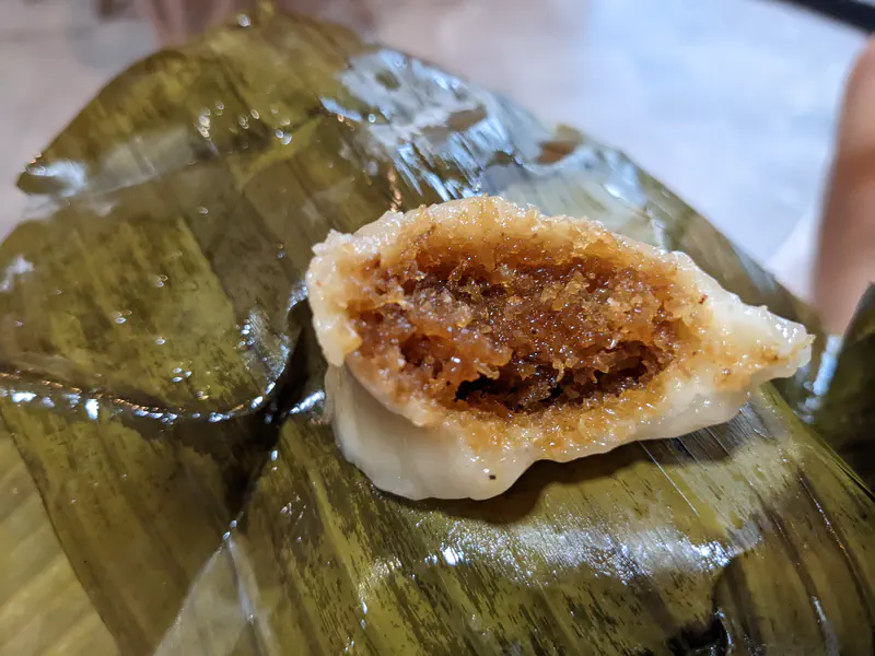 Close-up of a Thai dessert wrapped in banana leaf, showing a filling of sweet shredded coconut inside a glutinous rice shell.