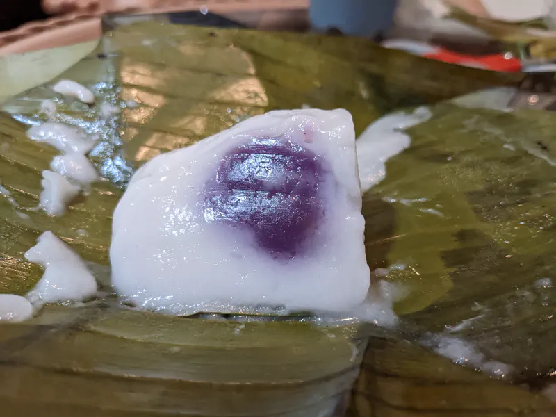 Close-up of a traditional Thai dessert with a white coconut layer and purple filling served on a banana leaf.