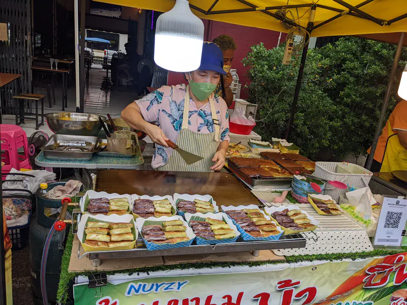 Street vendor cooking at a griddle with trays of fried pancakes and desserts displayed at a market stall.