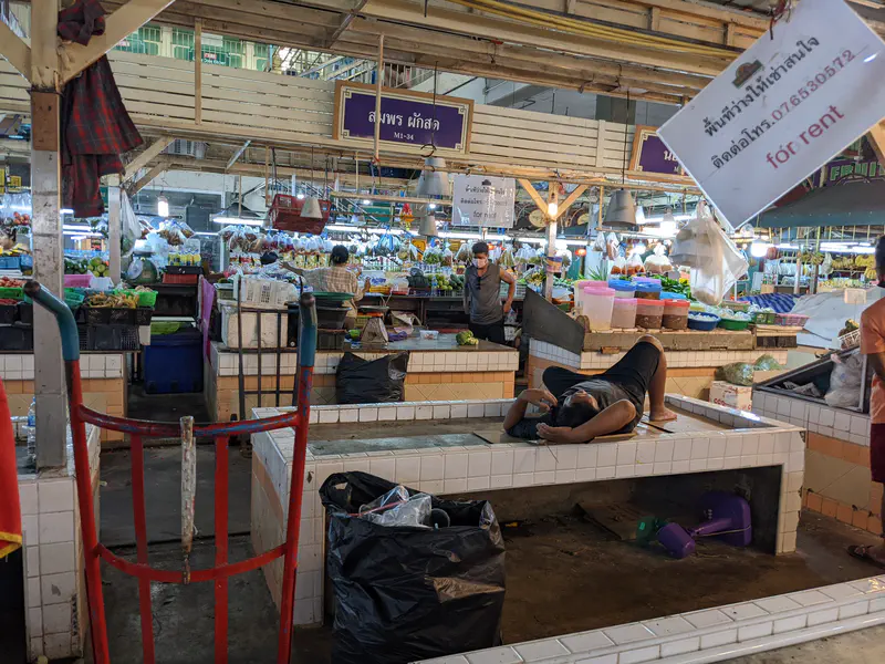 Man lying down and resting on an empty tiled vendor stall inside a busy fresh market.