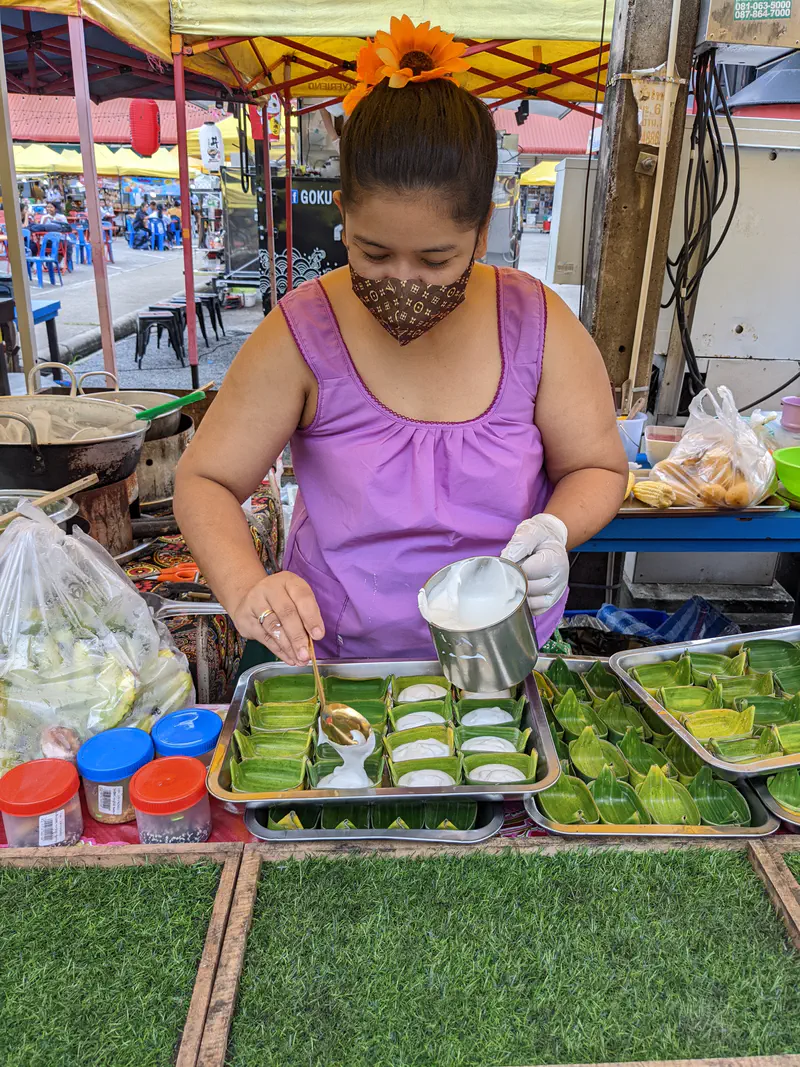 Woman in a purple top and mask pouring coconut cream into banana leaf cups to prepare traditional Thai desserts at a market stall.