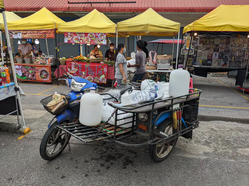 Motorbike with a sidecar loaded with bags and large plastic containers parked in front of yellow market stalls.