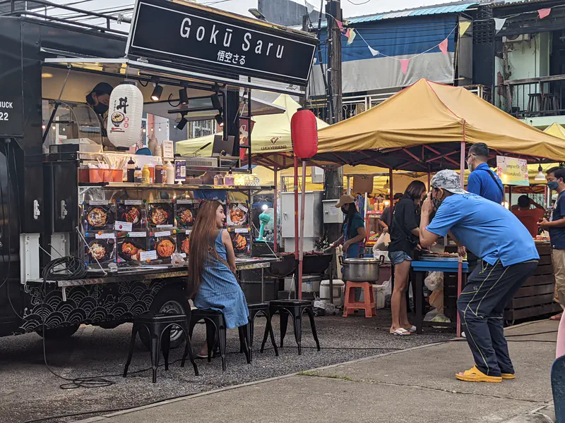Food truck with a display of Japanese dishes, a woman sitting in front posing for a photo taken by a man at a street market.