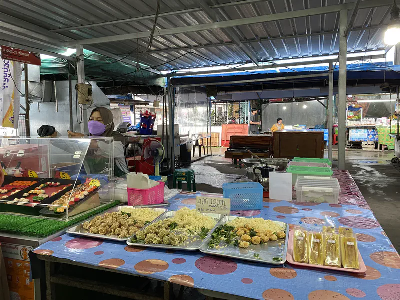 Market stall with trays of prepared food including fried snacks and sushi, with a vendor wearing a purple mask behind the counter.