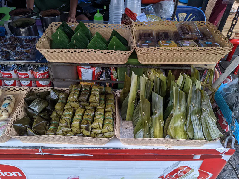 Market stall displaying traditional Thai desserts wrapped in banana leaves and plastic containers of sweets arranged in baskets.