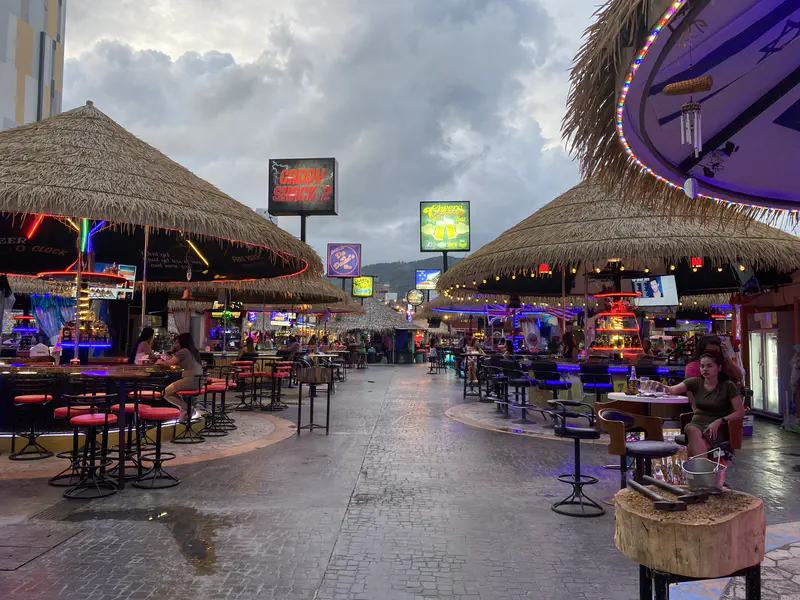 Outdoor nightlife scene with bars under thatched roofs, neon lights, and people sitting at tables as evening sets in.