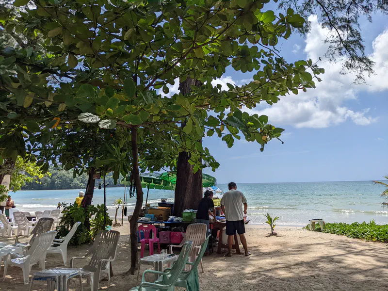 Beachfront scene with plastic chairs under shady trees, a small food stall, and a view of the calm sea.