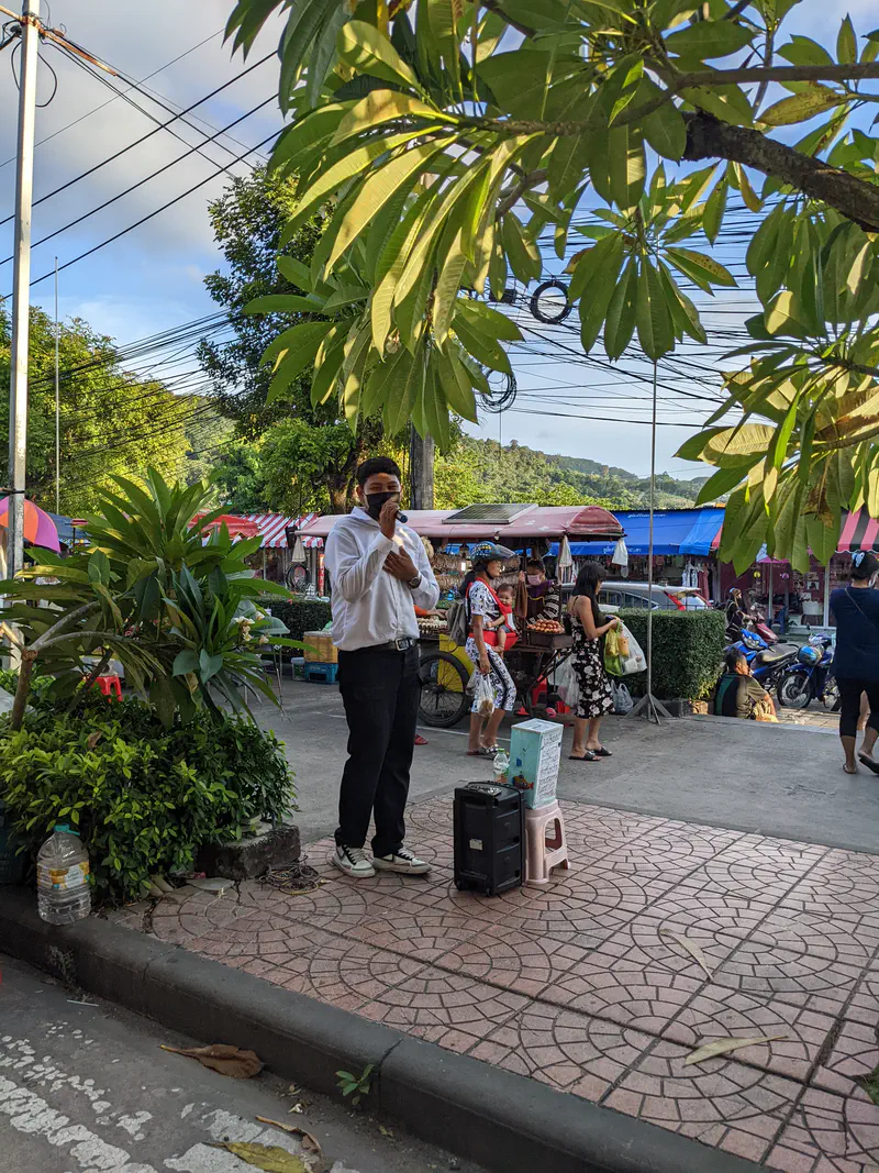 Man in white shirt speaking into a microphone with a speaker on the sidewalk, near a busy market area.