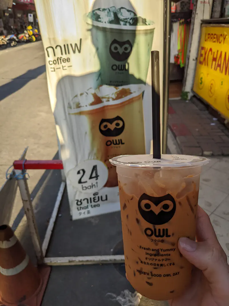 Hand holding a cup of iced Thai tea in front of a street sign advertising drinks for 24 baht.