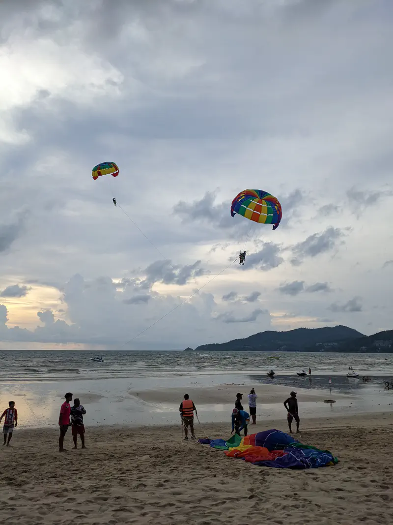 People parasailing with colorful parachutes over the ocean at a beach during cloudy sunset.