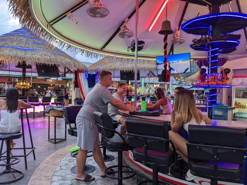 People sitting at a circular bar with neon lighting and a central pole inside a thatched-roof nightlife venue.