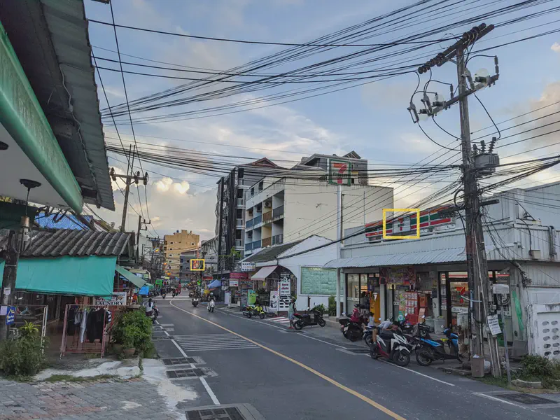Street scene with motorbikes, shops, and a 7-Eleven convenience store under a web of overhead power lines.