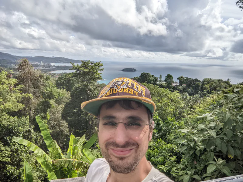 Man taking a selfie at a viewpoint with lush greenery, ocean, and coastal town in the background under a partly cloudy sky.