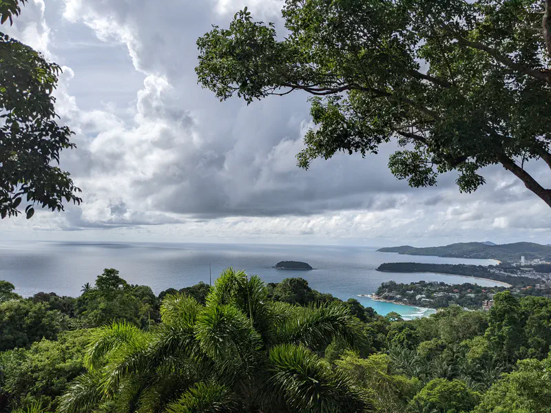 Scenic viewpoint overlooking the ocean, small islands, and a coastal town framed by lush green trees under a cloudy sky.