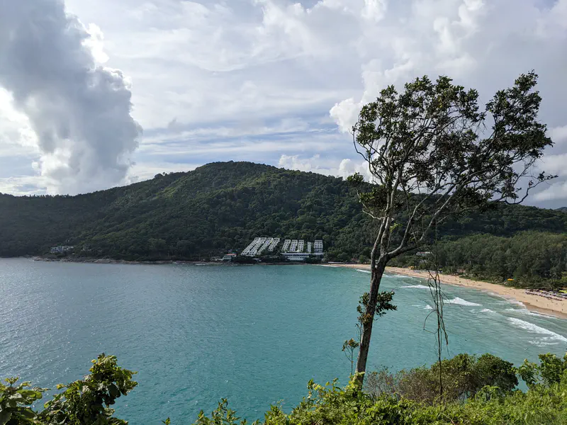 Scenic view of a turquoise bay with a sandy beach, forested hills, and buildings along the shore under a partly cloudy sky.