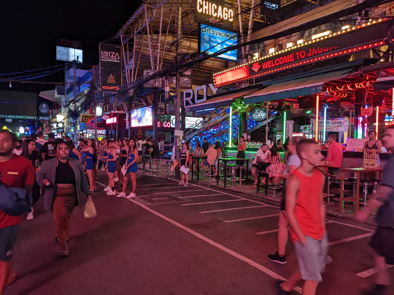 Nightlife scene on a busy street with neon lights, crowded bars, and people walking along the road in front of clubs.