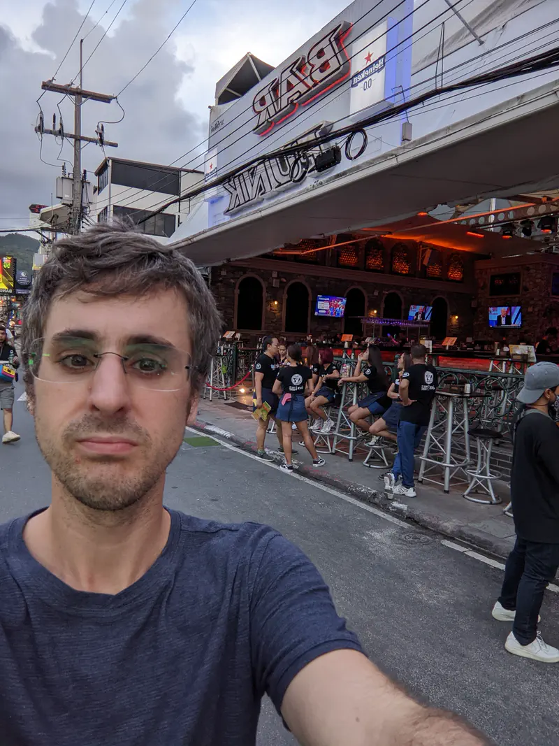 Man taking a selfie on a busy street in front of a bar with people sitting outside under a cloudy sky.