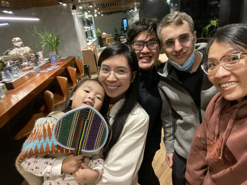 Group of five people smiling indoors, with a child holding a colorful fan.