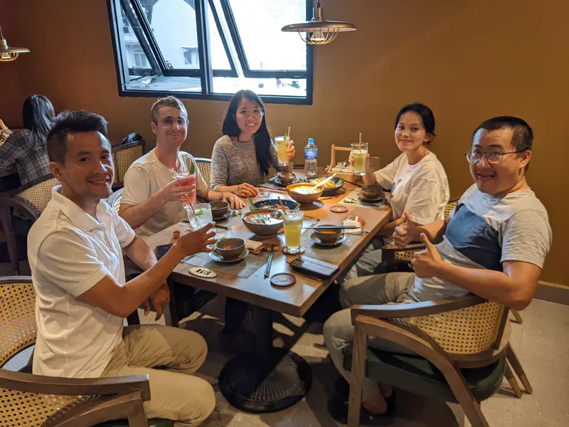 Group of five people sitting at a restaurant table with drinks and food, smiling at the camera.