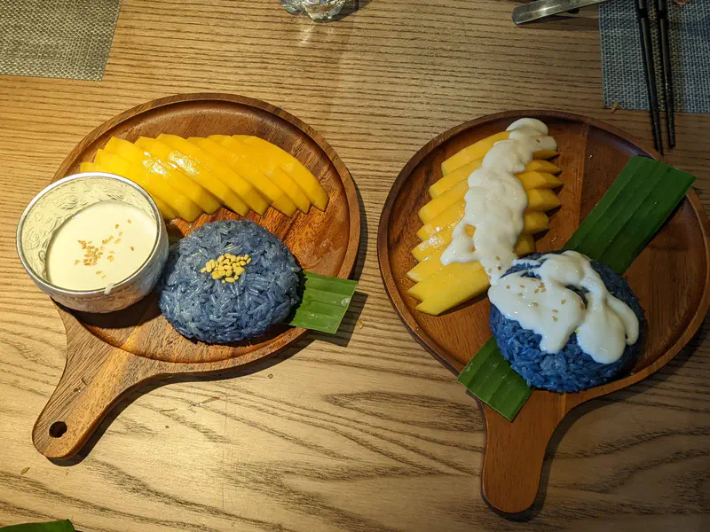 Plates of mango sticky rice with blue rice and coconut sauce on wooden trays.