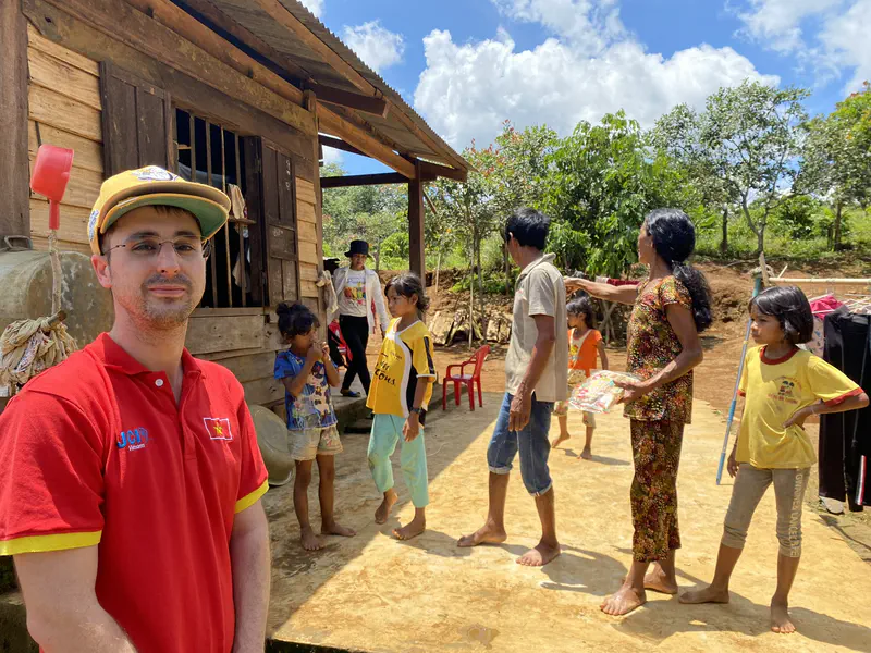 Man in a red shirt standing near a wooden house with several children and adults outside.