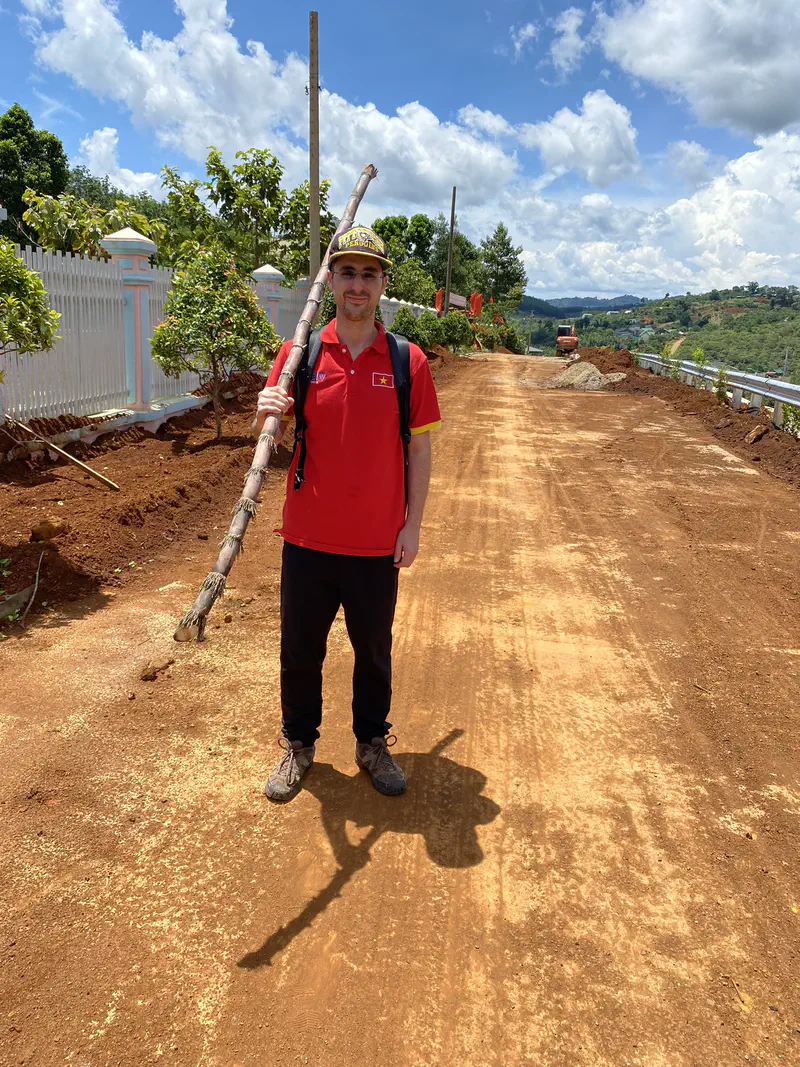 Man in a red shirt and cap holding a sugarcane on a dirt road with hills in the background.
