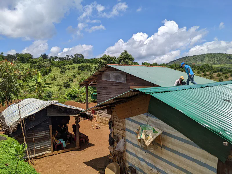 Two people working on the metal roof of a wooden house in a rural area with hills and trees.