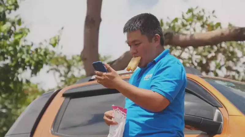 Man in a blue shirt holding a phone while eating bread near an orange car.
