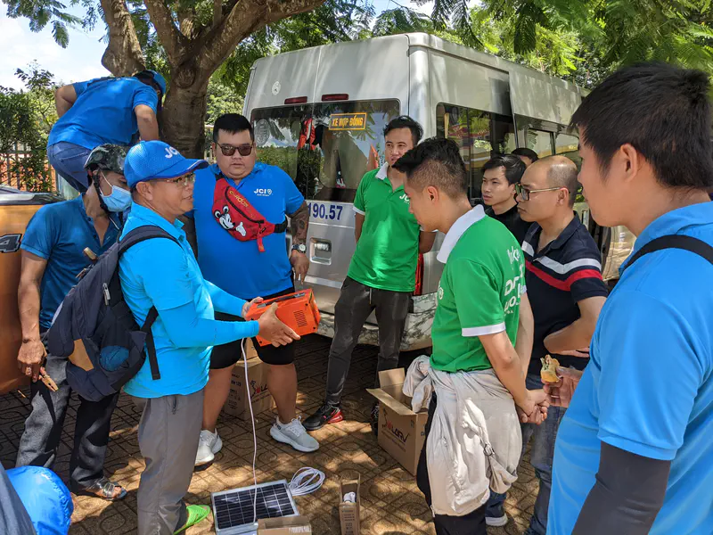 People gathered outdoors while a man demonstrates a small orange generator with solar panels.