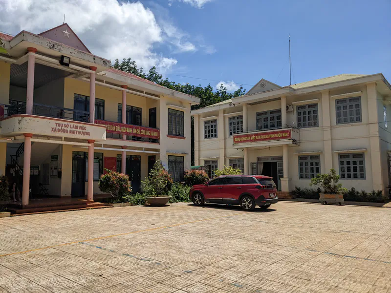 Two government buildings in Đồng Nai Thượng with banners and a red car parked outside.