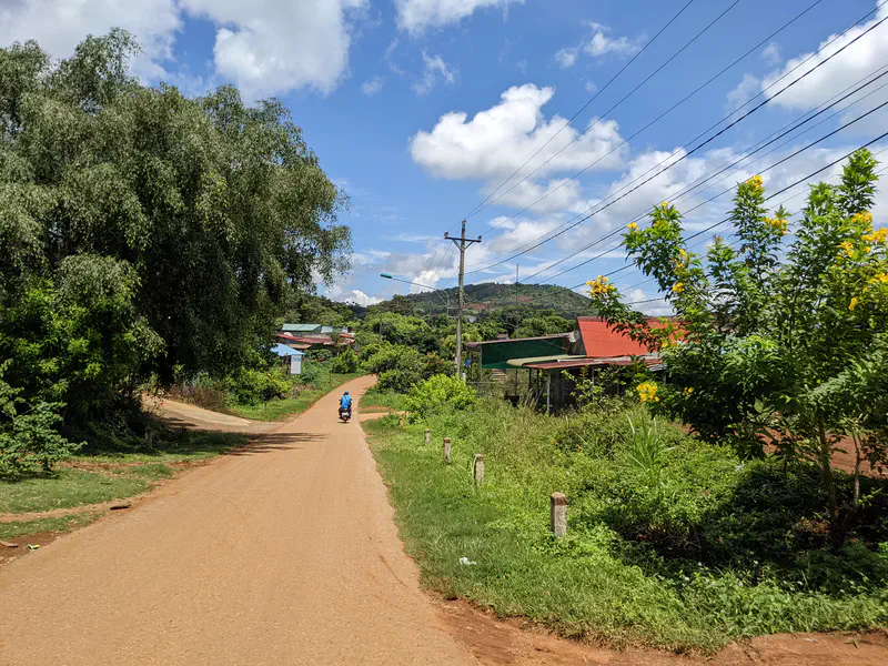 Rural dirt road with a person on a motorbike, trees, houses, and hills under a blue sky.