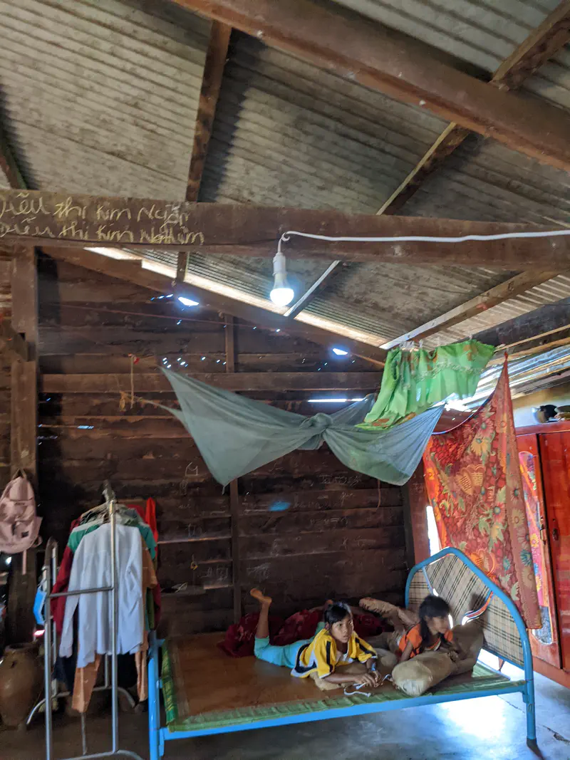 Two children lying on a bed under a mosquito net inside a wooden house with clothes and furniture.