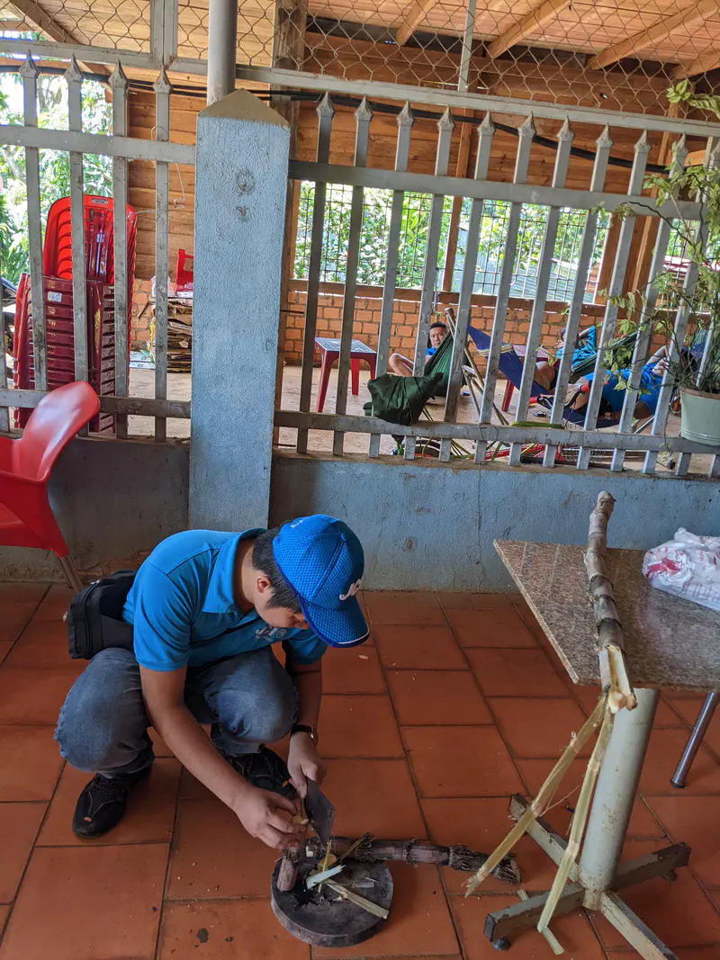 Man in a blue shirt and cap cutting sugarcane on a tiled floor with people resting in hammocks nearby.