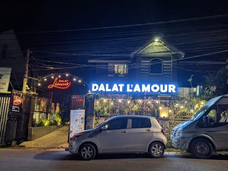 Night view of Dalat L’Amour restaurant with bright signs, lights, and parked cars outside.