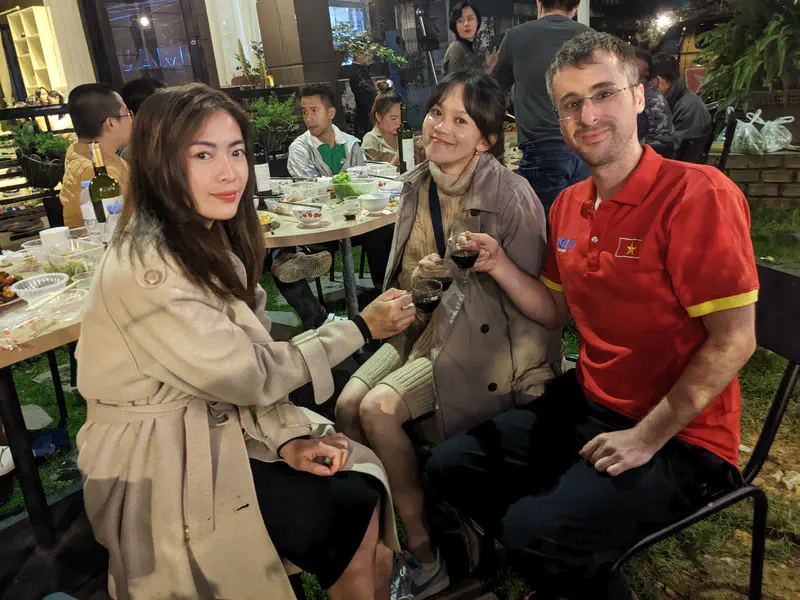 Three people sitting at an outdoor table raising glasses together during a dinner gathering.