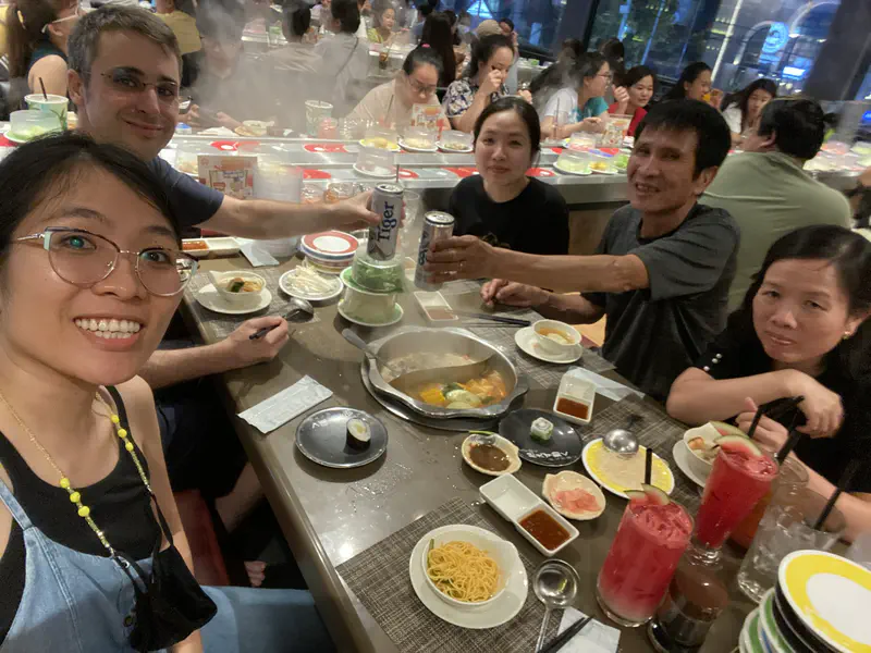 Group of people enjoying hot pot dinner together at a busy restaurant, with food, drinks, and steam rising from the pot.
