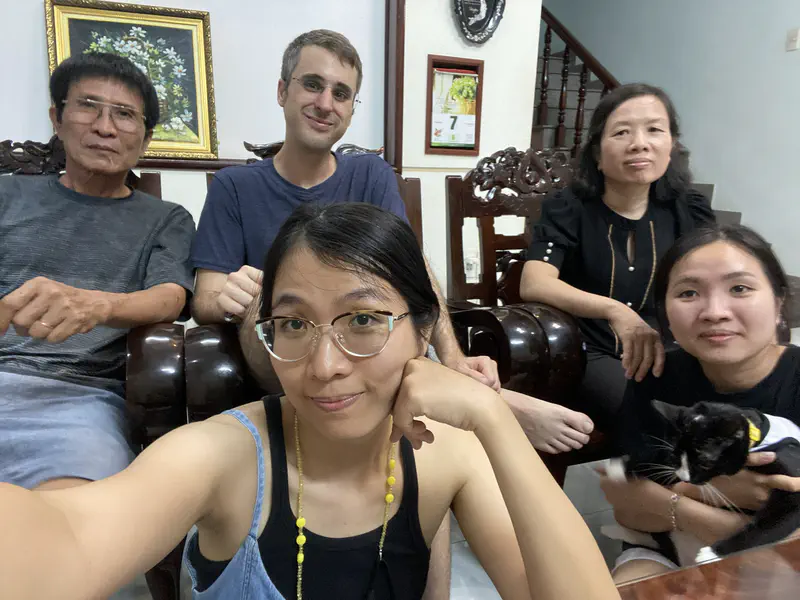 Five people sitting indoors on wooden chairs, with one person holding a black and white cat.